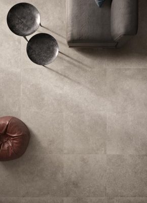 Viršutinis vaizdas į modernų svetainės interjerą su tamsios spalvos sofa, apvaliais metaliniais staliukais ir odiniu tamsiai rudu pufu. Grindys padengtos natūralaus akmens imitacijos pilkomis plytelėmis.