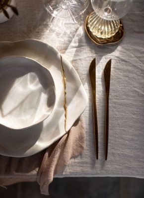 Elegantiškai padengtas stalas su baltos keramikos lėkštėmis, auksinio atspalvio stalo įrankiais ir krištolinėmis taurėmis ant tekstūrinės staltiesės.